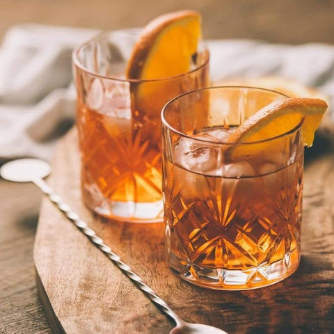 Two glasses of **Magic Hour Gamma-Aminobutyric Acid (GABA) Oolong Tea** garnished with orange slices sit on a wooden board. The amber liquid inside the glasses reflects warm light, showcasing their intricate glass patterns. A long-handled stirring spoon rests beside the glasses on the board, offering a calm nourishment to enhance the mood.