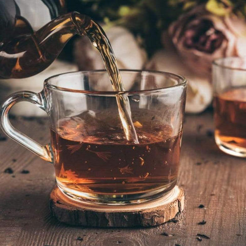 A glass teapot pours Magic Hour Gamma-Aminobutyric Acid (GABA) Oolong Tea into a clear glass cup placed on a wooden coaster. Another cup of tea is in the background, slightly out of focus, enhancing the calm nourishment of the rustic setting with muted tones and blurred flowers.