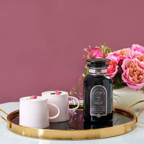 A bottle of Gypsy Rose Black Tea by Magic Hour sits on a round golden tray with a black base. Next to it are two white cups filled with tea, garnished with small rose petals. In the background is a bouquet of pink roses against a mauve wall.