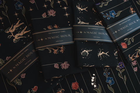 Four packaged Tea Ceremony Towels labeled "Magic Hour" lie on a decorated surface. Each towel features light gold and colored floral and celestial designs against a dark background. Two packages showcase constellations, while the other two highlight flowers.