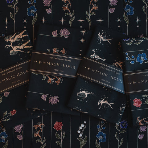 Four Tea Ceremony Towels are displayed on a dark, patterned surface. Each towel is wrapped with a band that reads "Tea Ceremony Towels by Magic Hour." These elegant pieces, perfect for your Magic Hour tea rituals, feature intricate designs of flowers, celestial bodies, and constellations.