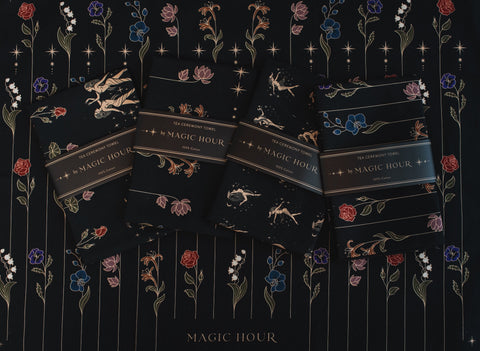 Four dark-colored packets of Magic Hour Tea Ceremony Towels are arranged in a row on a floral-patterned background. The packets feature small illustrations of celestial elements and flowers in shades of gold, white, and blue, capturing the enchanting essence of this organic loose leaf tea.