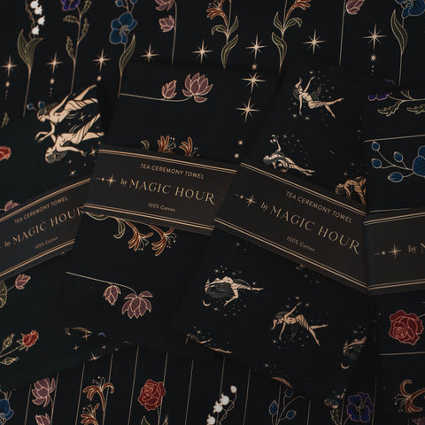 A collection of elegant Tea Ceremony Towels by "Magic Hour," featuring intricate floral and celestial designs in gold, blue, and red on a black background. Each towel is labeled "100% Cotton." They are artistically arranged, showcasing their elaborate patterns and perfect for your loose leaf tea rituals.