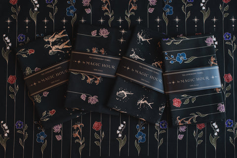 Four black tea ceremony towels labeled "Tea Ceremony Towels" by Magic Hour are laid out on a floral-patterned fabric. Each towel features intricate designs depicting constellations, floral motifs, and starry elements, showcasing a celestial theme. Enjoy with your favorite organic loose leaf tea for a truly magical experience.