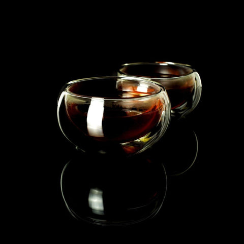 Two glass teacups filled with dark liquid, possibly Sin Eraser™ : Puerh Tea by Magic Hour, are placed on a glossy black surface. The cups have a double-walled design, creating a clear and elegant appearance, with reflections visible beneath them. The background is completely black.