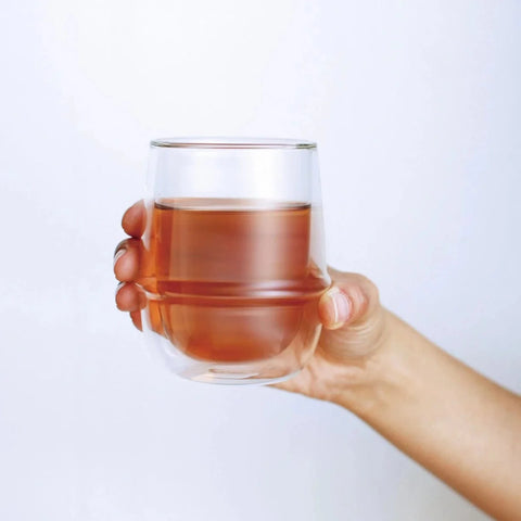 A hand is holding a Kinto Double-Walled Iced Tea Glass: 350mL filled with a light brown liquid against a plain white background. The glass is rounded and transparent, showcasing the Magic Hour Organic Tea inside clearly.