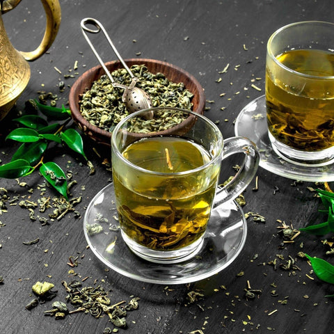 Two glass cups of Magic Hour Mao Jian (Emerald) Green Tea of Good Fortune on saucers sit on a dark surface, surrounded by loose green tea leaves. A wooden bowl filled with more organic tea leaves and a tea strainer are nearby. Green tea leaves and a brass teapot add to the cozy, relaxed atmosphere.