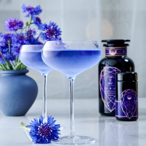 Two elegant cocktail glasses filled with a vibrant blue drink sit on a white surface, reminiscent of the soothing Sapphire - Apple-Plum-Spice White Tea by Magic Hour. A purple flower is placed beside one glass. In the background, there is a vase with similar purple flowers and two dark bottles adorned with intricate purple designs.