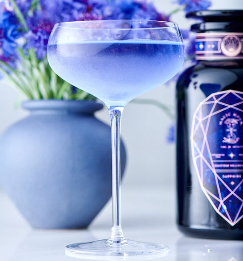 A delicate cocktail glass filled with lavender-colored Sapphire - Apple-Plum-Spice White Tea from Magic Hour is in the foreground. Behind it, there's a blue vase containing purple flowers and a stylish dark bottle with a geometric design on the label, all set against a light background.