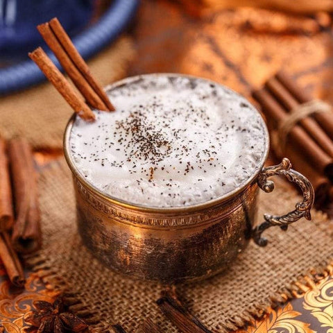 A vintage metal cup filled with frothy white beverage topped with ground spices and garnished with cinnamon sticks. It's placed on a textured mat surrounded by loose leaf tea, cinnamon sticks, and star anise, set against a warm, rustic background. The scene features Magic Hour's Queen of the South: Delicious Cocoa Detox Tea.