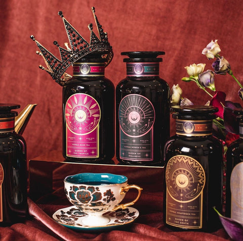 A luxurious display of three ornate tea containers featuring Queen of Wellness: Women's Hormone Balancing Tea for PMS, Healthy Cycles & Menopause by Magic Hour with regal labels, set against a rich, burgundy backdrop. A decorative crown rests atop one container, and a beautifully detailed teacup and saucer sit nearby. Delicate flowers add a touch of elegance to the scene.