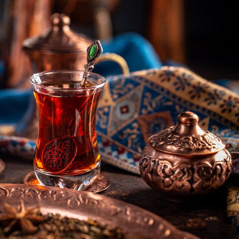 A glass of Sin Eraser™: Puerh Tea by Magic Hour with a decorative metal spoon sits on a copper saucer. The background features an intricate patterned cloth and an ornate copper container with a lid. The scene is richly colored and exudes a warm, traditional atmosphere.