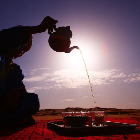 A person is pouring Sin Eraser™ : Puerh Tea from Magic Hour from a teapot into glasses on a tray set on a red cloth in a desert. The silhouetted figure is against a setting sun, creating a serene atmosphere with the sky transitioning from light to dark.