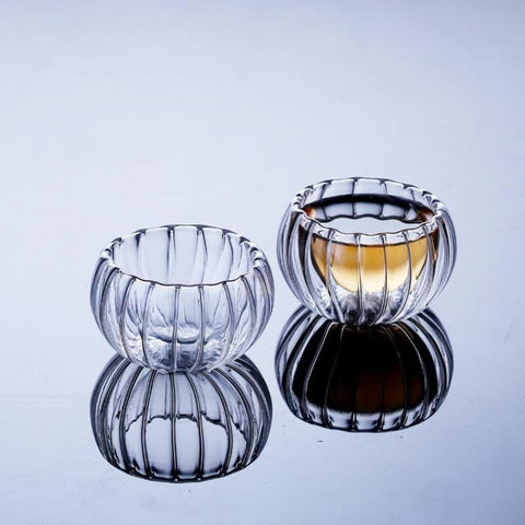 Two ribbed, transparent glass bowls sit on a reflective surface. One is empty, while the other, reminiscent of the Double-Walled Rippled Tea Cup, is half-filled with a golden liquid. The reflection of the bowls is visible beneath them.