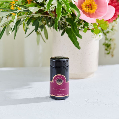 A jar of Raspberry Earl White Tea from Club Magic Hour sits on a white table with a colorful flower arrangement in the background. The jar has a dark lid and a pink label. Natural light illuminates the scene, creating the perfect setting for your next cup of Organic Tea.