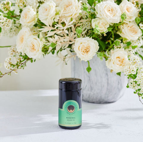 A jar of organic honey labeled "Renewal" sits on a white table alongside a tin of Renewal: Peach-Goji-Rose Oolong Tea by Magic Hour, in front of a large bouquet of white and light pink roses and other white flowers in a gray vase. The scene is bright and airy, suggesting a calm and refreshing atmosphere.