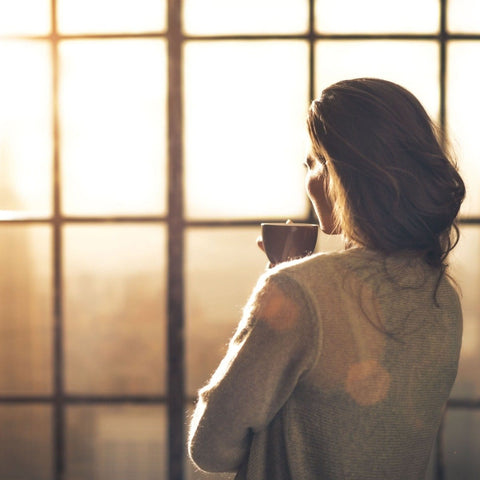 A person with long hair is standing indoors, holding a mug of Rise & Shine Black Tea from Club Magic Hour, and looking out of a large window at a bright morning sun. They are dressed in a cozy sweater, and the atmosphere is warm with a soft, golden light filtering through the window.