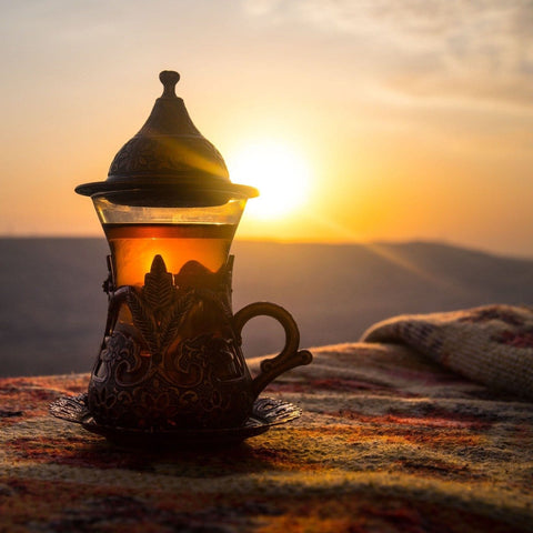 A decorative glass teacup with an ornate metal handle and lid sits on a colorful woven blanket. The cup contains Rise & Shine Black Tea by Club Magic Hour, illuminated by the warm glow of a Magic Hour Tea sunset in the background, casting a serene and tranquil ambiance.