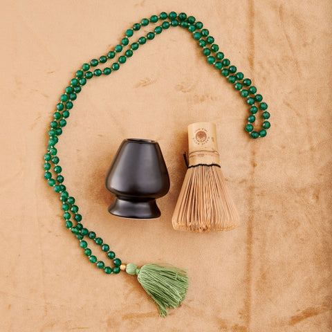 An arrangement on a beige surface features a green beaded necklace with a green tassel, a black ceramic tea bowl, and the Matte Black Chasen - Matcha Whisk Stand holding the bamboo whisk used in Japanese tea ceremonies. The necklace is arranged in a loose circle around the bowl and whisk stand.