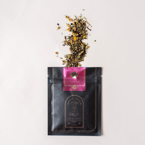 A black and gold Magic Hour tea packet labeled "Symbeeosis: Beautifying Immunitea for the Queen Bee" lies flat on a white surface. The packet is opened with loose leaf tea and flower petals spilling out onto the surface above the packet, offering a glimpse of this exquisite organic tea blend.