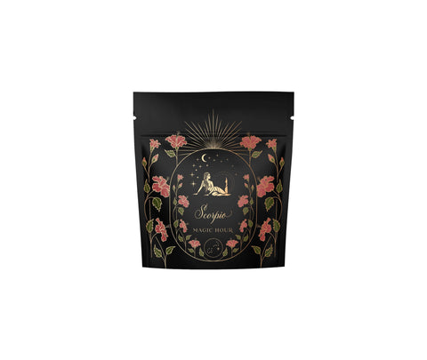 A black package of Scorpio Tea for Sensual Brilliance by Magic Hour features a celestial design with a crescent moon, stars, and a seated figure gazing up. The packaging is adorned with pink flowers and golden decorative elements, emphasizing the premium quality of this organic tea.