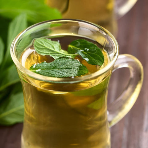 A clear glass cup filled with hot Organic Spearmint Herbal Tisane garnished with fresh mint leaves. Another glass with the same beverage is blurred in the background. The setting includes some more organic mint leaves on a wooden surface beside the cups.