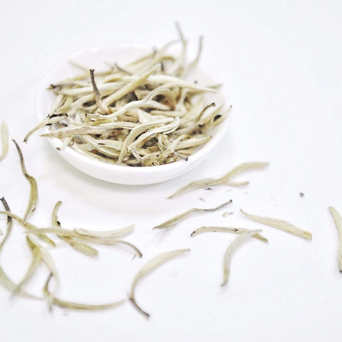 A small white bowl filled with Silver Moon White Tea leaves from Club Magic Hour is placed on a clean, white surface. Several thin, elongated leaves are scattered around the bowl, creating a minimalist and clean visual. The pale color of the Silver Moon White Tea adds to the serene aesthetic.