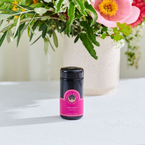 A vibrant bouquet with various flowers in a white vase sits on a white surface. In the foreground, there is a black jar with a pink label that reads "Magic Hour Soulmate: Chocolate-Raspberry-Rose Black Tea for Finding & Celebrating Love" in front of the vase. The background is softly blurred, highlighting the jar and flowers.