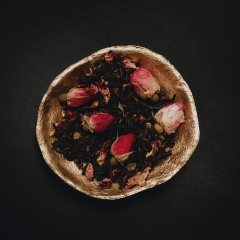 A golden bowl filled with a blend of Soulmate: Chocolate-Raspberry-Rose Black Tea for Finding & Celebrating Love by Magic Hour, set against a dark background. The delicate pink and white rose buds add a touch of color to the deep brown tones of the organic tea leaves, creating an enchanting scene worthy of a magic hour.