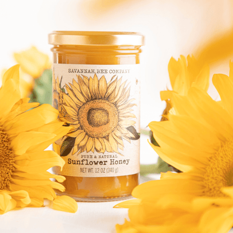 A jar of Savannah Bee Co. Honey - Sunflower is surrounded by vibrant yellow sunflowers. The label features an illustration of a sunflower and contains 12 ounces (340 grams) of pure and natural sunflower honey, sourced from the rich fields of Ukraine and Romania.