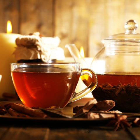 A clear glass cup filled with dark, Magic Hour's Symbeeosis: Beautifying Immunitea for the Queen Bee is placed next to a glass teapot on a wooden surface. Surrounding the tea set are cinnamon sticks, star anise, and a lit candle in the background, creating a cozy ambiance.