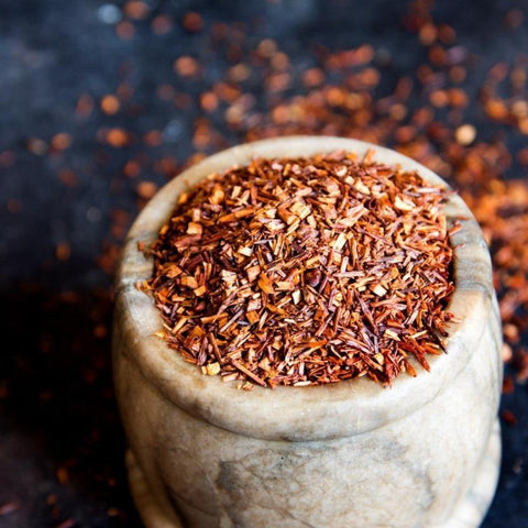 A rustic stone mortar is filled to the brim with finely shredded, reddish-brown, organic Queen of the Harvest : Herbal Relaxation Tea by Magic Hour. The dark background is scattered with additional loose leaf tea, highlighting the vibrant tone and texture of the rooibos.