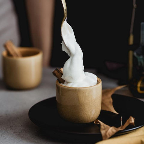 A spoon is lifting a swirl of whipped cream from a small beige cup. The cup sits on a black saucer with a cinnamon stick, dried leaves, and hints of loose leaf tea nearby. Another similar cup is in the background. The setting has a warm and cozy atmosphere, reminiscent of Magic Hour's Queen of the Harvest: Herbal Relaxation Tea moments.