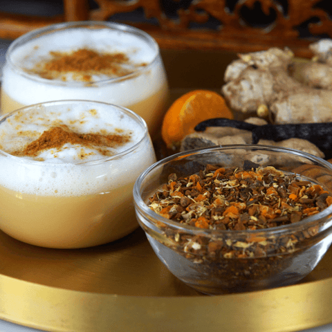 Two glass cups filled with frothy, spiced beverages are on a golden tray, each topped with ground cinnamon. Beside the cups is a bowl of mixed spices and scattered ginger roots. A whole nutmeg and a piece of turmeric complete the scene, perfect for an organic tea experience with Magic Hour's Queen of the Harvest: Herbal Relaxation Tea.