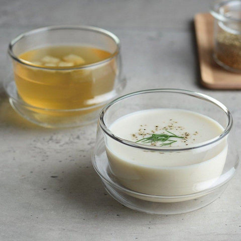 A table displays two glass bowls with handles. The foreground bowl is filled with creamy white soup garnished with herbs and black pepper. The background bowl contains clear broth with visible ingredients. A wooden tray, adorned with spices and a delicate cup of Kinto Double-Walled Teacups, is partially visible.