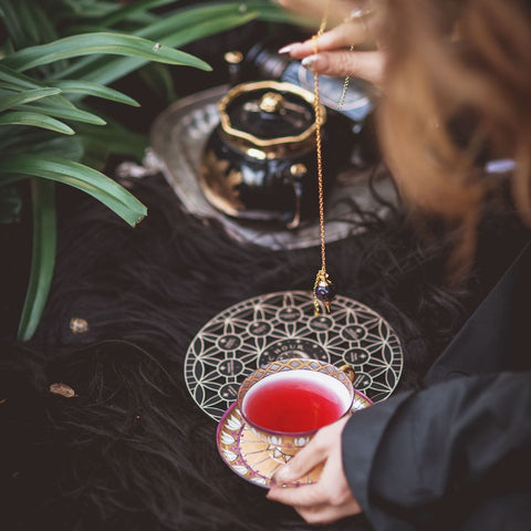 A person holding a teacup filled with red liquid in one hand and a pendulum in the other, over a decorative object on the ground. A black teapot of organic tea on a silver tray and green foliage are visible in the background. The product being showcased is the Crystal Grid: Flower of Life Tea Ceremony Altar by Alibaba.